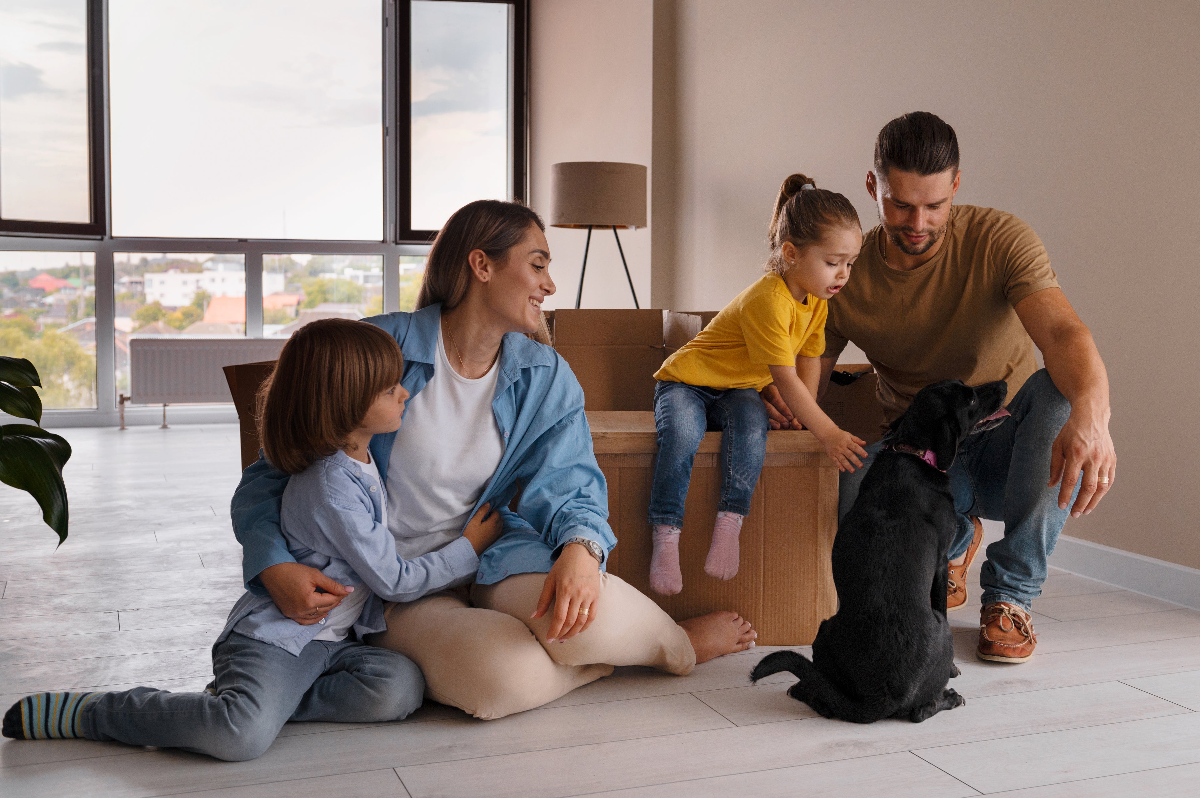 A happy family with two young children and a black dog in what appears to be a new or recently moved-into home. The parents and children are interacting with the dog, suggesting a comfortable and safe environment. The room is light-filled with large windows in the background.