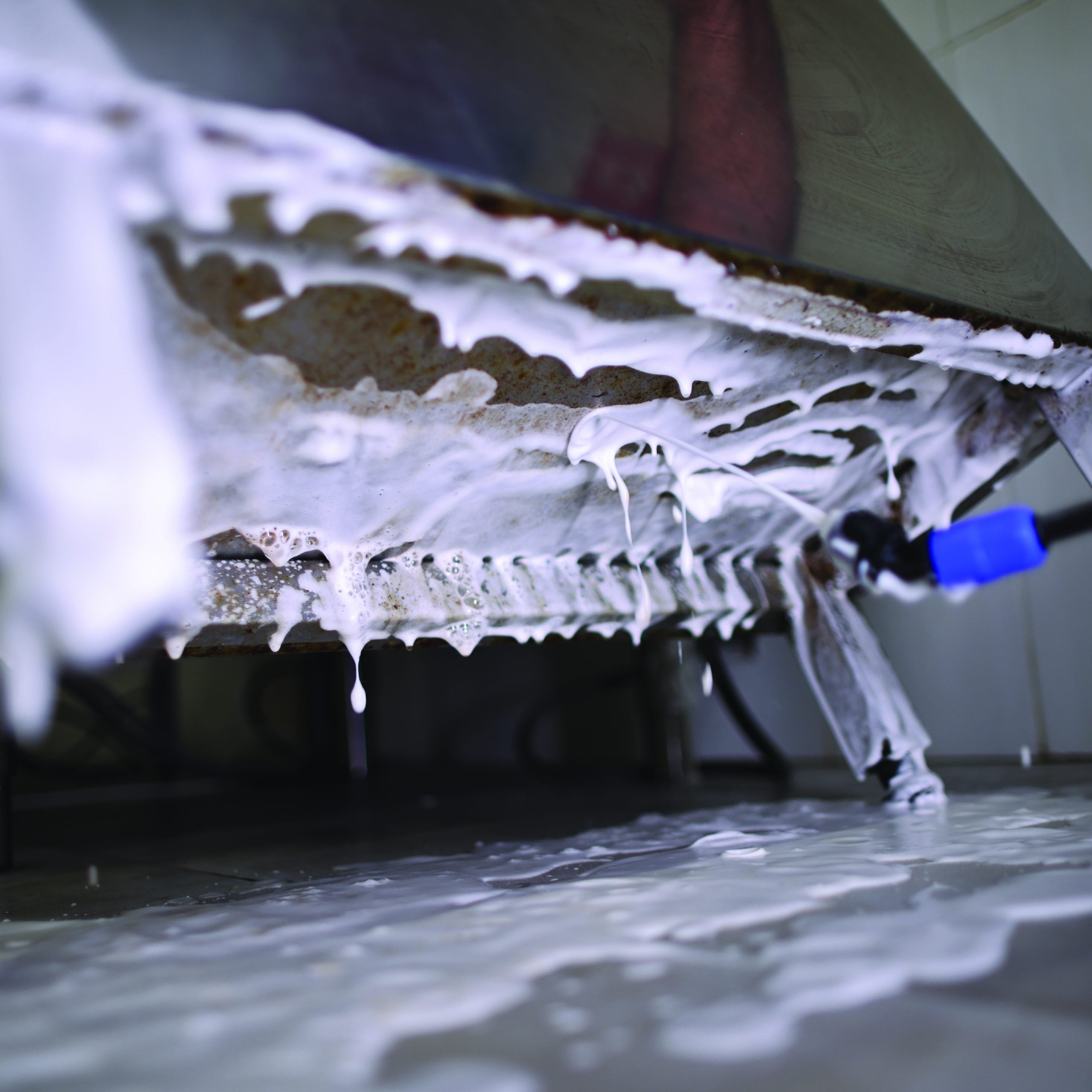 Close-up of commercial kitchen equipment being deep cleaned with foam disinfectant spray by a pest control professional using a blue-handled sprayer.